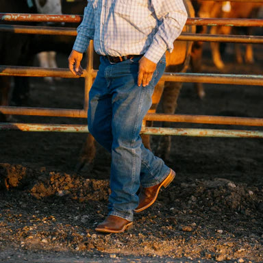 Man in a plaid shirt and jeans walking in a barn with cows in the background