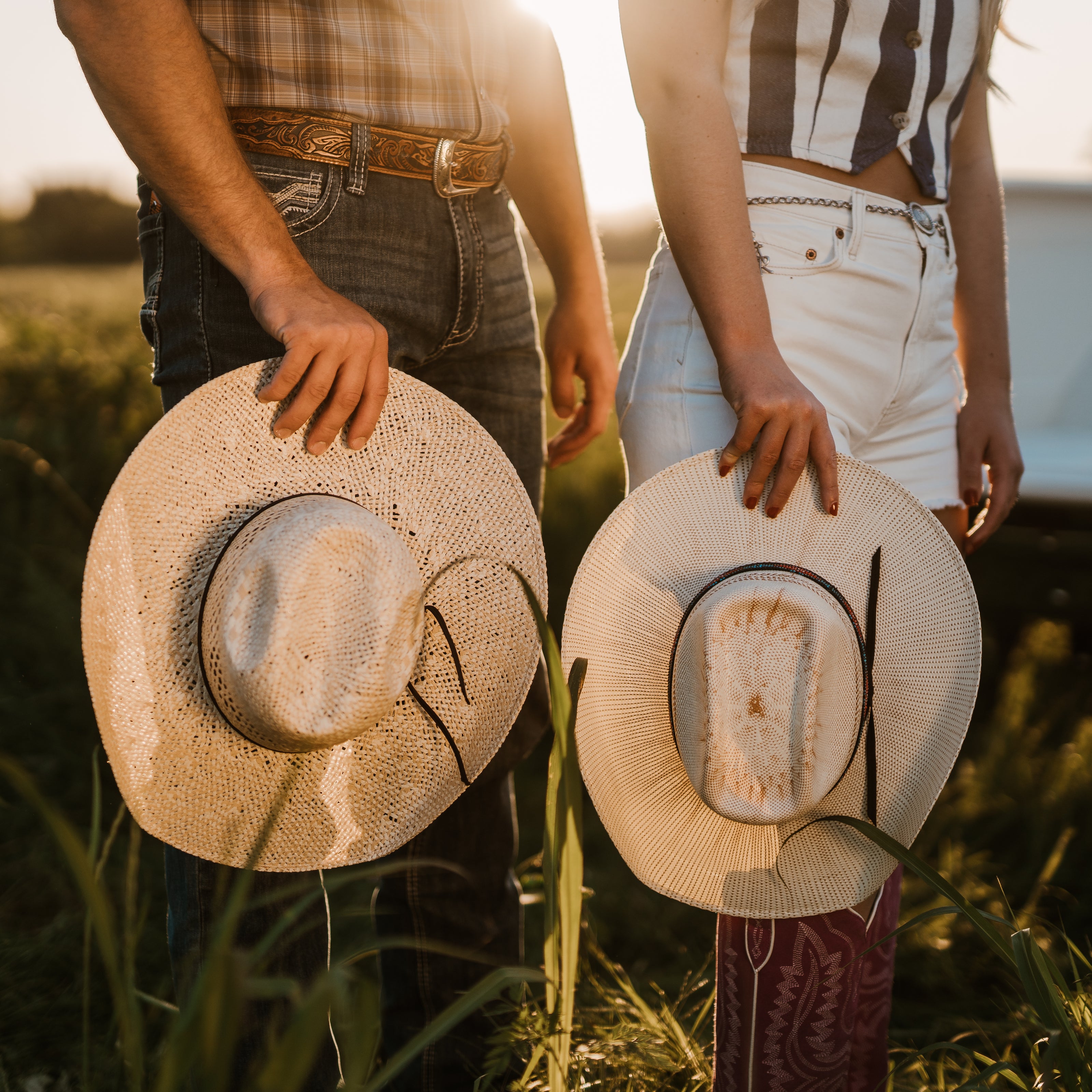 Two people in a field holding straw hats, wearing casual clothing.
