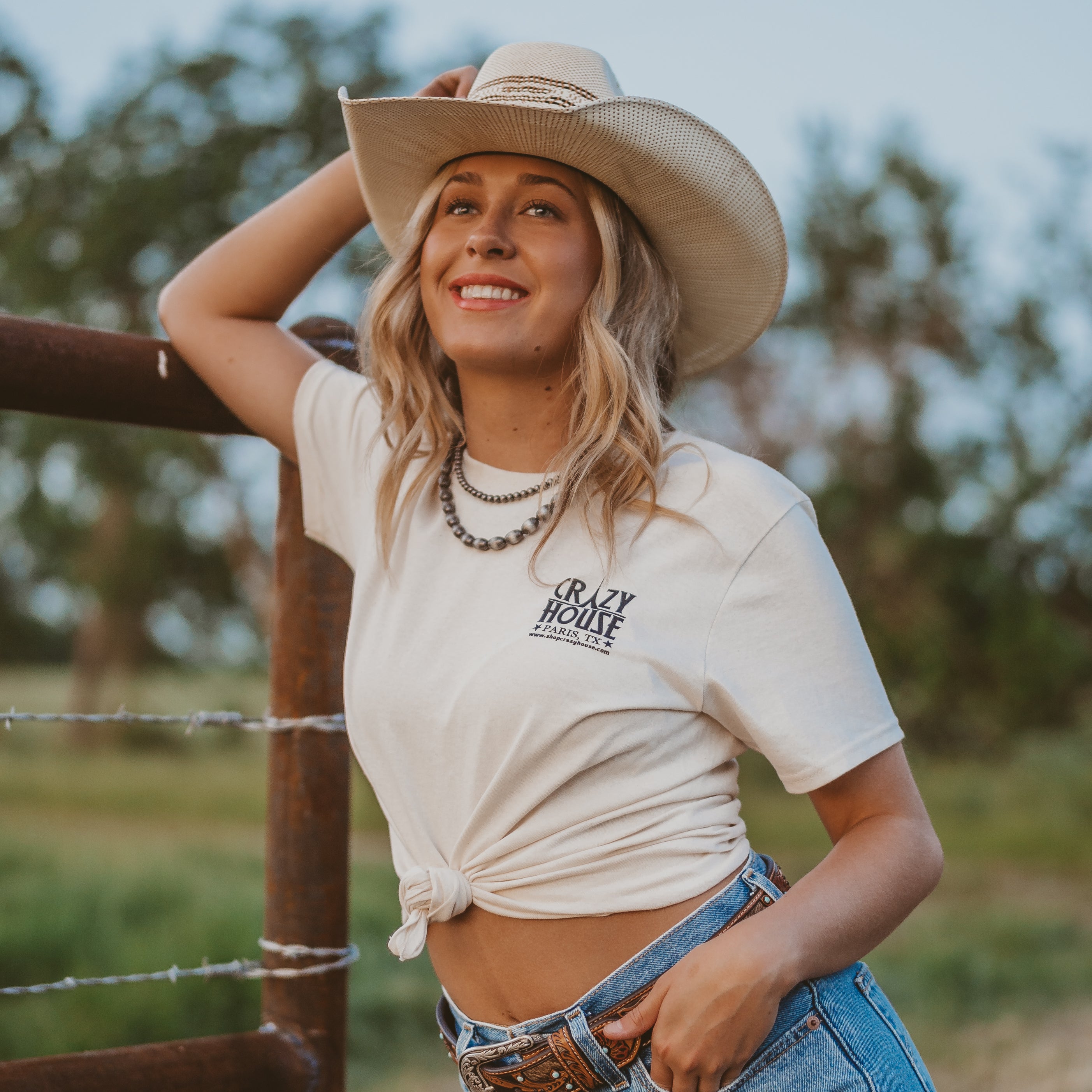 Woman in a cowboy hat and casual outfit standing by a fence in a natural setting