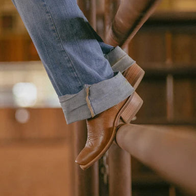 Person wearing brown leather shoes and rolled-up jeans sitting on a wooden stool.