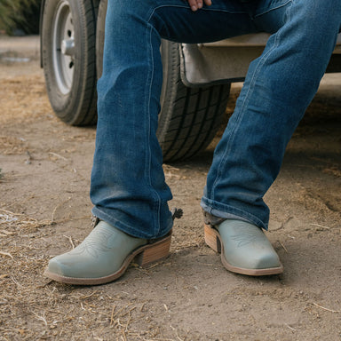 Person wearing blue jeans and light blue shoes standing on a dirt surface with a vehicle in the background.