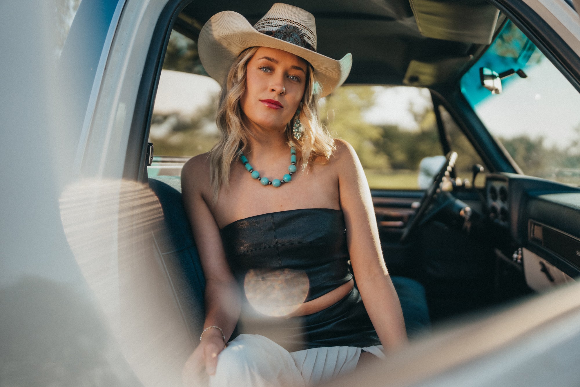 Woman sitting in a car wearing a black dress, white hat, and turquoise necklace.