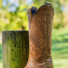 Brown cowboy boot with intricate designs leaning against a wooden post outdoors.